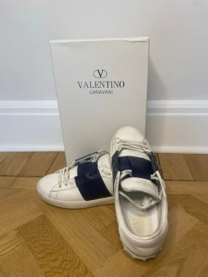 Valentino Garavani vita sneakers - Snygga vita sneakers från Valentino Garavani med bred marinblå detalj över snörningen. Skorna har klassisk rund tå, snörning och är tillverkade i skinn. Perfekta för dig som vill ha en stilren och exklusiv look. Storlek 42