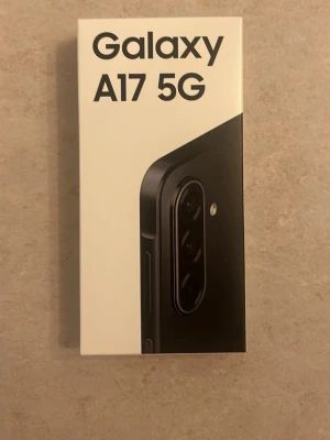 Samsung Galaxy A17 5G svart helt ny - Säljer en Samsung Galaxy A17 5G i svart. Telefonen har en modern design med rundade hörn och flera kameror på baksidan. Lådan är vit med tydlig logga och bild på mobilen. Perfekt för dig som vill ha en snabb och snygg smartphone med 5G-stöd.