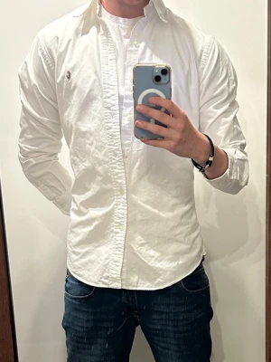 Vit skjorta från Ralph Lauren - Klassisk vit skjorta från Ralph Lauren med knappstängning och broderad logga på bröstet. Skjortan har lång ärm, normal passform och är tillverkad i bomull. Perfekt till jeans eller chinos för en clean och stilren look.
