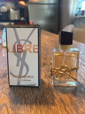 Yves Saint Laurent Libre EdP 50ml - Yves Saint Laurent Libre Eau de Parfum i en elegant, rektangulär glasflaska med gulddetaljer och svart lock. Flaskan har en lyxig design med YSL-loggan i guld runt flaskan. Doften är modern och ikonisk, perfekt för dig som gillar exklusiva parfymer. ca 45 ml kvar av 50 ml. Köpt för 1400 kr.