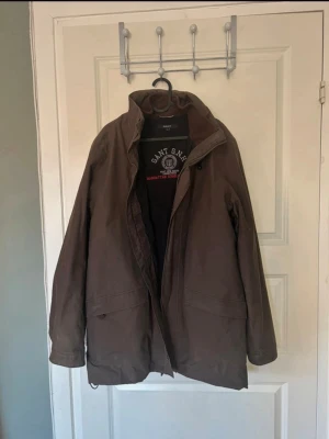 Brun vindjacka från Gant  - Säljer en brun vindjacka från Gant, modell Manhattan Double Jacket. Jackan har hög krage, dragkedja och tryckknappar framtill samt två fickor. Materialet är slitstarkt och jackan har en klassisk, rak passform med diskret logga på insidan.