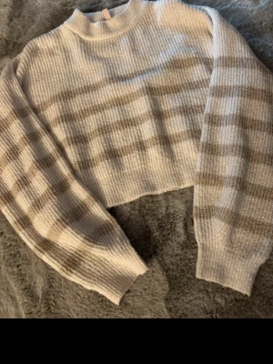 Beige-randig stickad tröja Nelly - Mysig stickad tröja från Nelly i beige och ljusgrått med breda horisontella ränder. Tröjan har croppad passform, ribbstickade muddar och rund hals. Perfekt för lager på lager och chill stil.