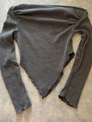 Grå ribbad långärmad topp - Trendig grå ribbad topp med lång ärm och asymmetrisk, spetsig nederkant. Perfekt för dig som gillar en edgy och unik stil. Materialet är mjukt och stretchigt, vilket gör den bekväm att bära hela dagen.