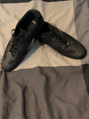 Svarta sneakers från HUGO BOSS - Stilrena svarta sneakers från BOSS med klassisk låg profil och snörning. Skorna har diskret logga på sidan och är gjorda i en mix av läder och textil för en modern look. Perfekta för dig som gillar minimalistisk och clean stil.