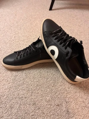 Svarta sneakers från Oliver Cabell - Svarta sneakers i läder från Oliver Cabell med vit sula och vit detalj på sidan. Skorna har svarta skosnören med märkesnamnet tryckt på och en vit hälflik. Snygg och stilren design som passar till det mesta.