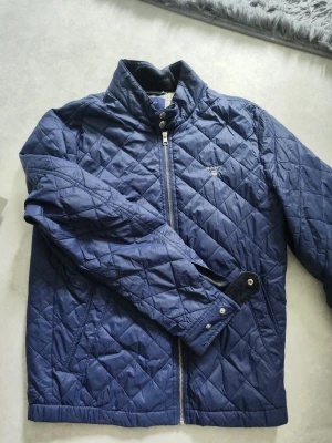Marinblå quiltad jacka från Gant - Snygg marinblå quiltad jacka från Gant med dragkedja och tryckknappar vid kragen. Jackan har ett klassiskt rutmönster och broderad logga på bröstet. Perfekt för dig som vill ha en stilren och tidlös look.