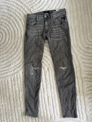 Gråa jeans från Replay W31 L34 - Säljer ett par grå jeans från Replay med slitningar på benen och klassiska fem fickor. Jeansen har dragkedjegylf och svarta sömmar som kontrast. Perfekt för dig som gillar en avslappnad men trendig look. Storlek W31 L34