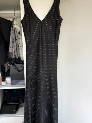 Svart lång prickig klänning - Svart långklänning med vita prickar och v-ringning. Ärmlös modell med smickrande passform och lätt utställd nederdel. Perfekt för dig som gillar en stilren och klassisk look.