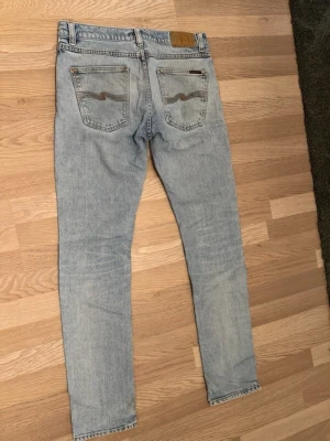 Ljusblå jeans från Nudie - Säljer ett par ljusblå skinny jeans från Nudie med snygga slitningar på ena knät. Jeansen har Nudies signaturbrodyr på bakfickorna och patch i läder bak i midjan. 