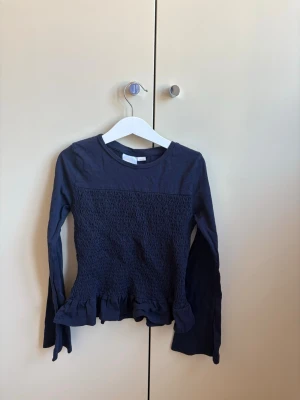Mörkblå långärmad topp med volang - Mörkblå långärmad topp med smockad framsida och volangdetalj nedtill. Rund halsringning och mjukt bomullsmaterial. Perfekt för en trendig och bekväm look.