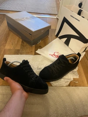 Svarta sneakers från Christian Louboutin - Stilrena svarta sneakers från Christian Louboutin i mocka med klassisk låg siluett. Skorna har svarta skosnören, svart sula och diskreta detaljer. Insidan är ljusbeige och märket syns på både dustbag och kartong. Storlek 41 passar 42