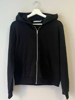 Svart hoodie med dragkedja från 157 - Svart hoodie från 157 med dragkedja framtill och stor huva. Klassisk modell med känguruficka och ribbade muddar vid ärmslut och nederkant. Perfekt för en avslappnad och stilren look. Tillverkad i mjukt material för extra komfort.