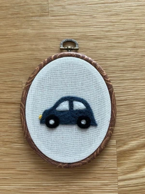 Broderad tavla med bilmotiv - Handgjord oval tavla med en nålfiltad mörkblå bil på ljus bakgrund, inramad i en oval ram. Perfekt som unik väggdekoration till barnrum och att matcha med andra motiv. Endast tavlan med bil som säljs. 