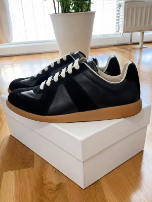 Maison Margiela Gats - Stilrena svarta sneakers från Maison Margiela med beige sula och vita snören. Skorna har en klassisk låg siluett och är tillverkade i en mix av läder och mocka. Perfekta för dig som gillar minimalistisk och tidlös design.