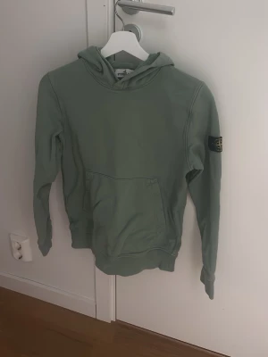 Grön hoodie från Stone Island - Snygg grön hoodie från Stone Island med klassisk patch på ärmen. Hoodien har en stor känguruficka framtill och huva med dragsko. Tillverkad i mjukt bomullsmaterial som är skönt mot huden. Perfekt för en avslappnad streetwear-look.