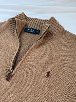 Polo Ralph Lauren Half Zip Sweater - Klassisk beige stickad tröja från Polo Ralph Lauren med halv dragkedja och ribbad krage. Tröjan har långa ärmar, ribbade muddar och den ikoniska broderade loggan på bröstet. Perfekt för lager på lager och stilren look.