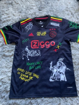 Ajax x Bob Marley svart matchtröja - Oanvänd fast utan prislapp. Säljer en svart Ajax x Bob Marley matchtröja från Adidas med färgglada detaljer i rött, gult och grönt. Tröjan har tryck med texter, musiknoter och illustrationer samt Ajax-logga och Ziggo-sponsor. Materialet är lätt och andas, perfekt för fotboll eller streetwear.