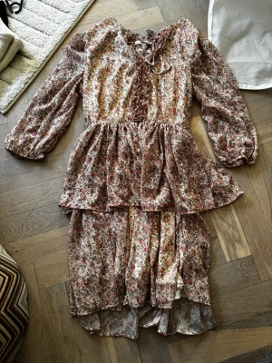 Blommig volangklänning med snörning - Romantisk blommig klänning med volanger och snörning framtill. Långärmad modell med v-ringning och mjukt fall. Perfekt för dig som gillar vintagekänsla och feminina detaljer.