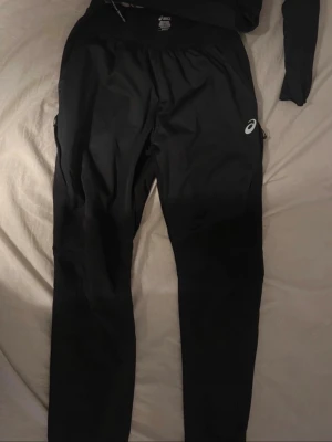 Svarta träningsbyxor från Asics - Svarta träningsbyxor från Asics med smal passform och elastisk midja. Byxorna har dragkedjor vid bensluten och en liten logga på låret. Perfekta för löpning eller gym.