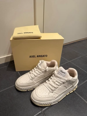 Vita sneakers från Axel Arigato - Säljer ett par stilrena vita sneakers från Axel Arigato, skorna har en diskret logga i guld på sidan. Använt 1 gång, man får med orginalkartong samt dustbag och påse.  Nypris 3500! Säljer för 2500