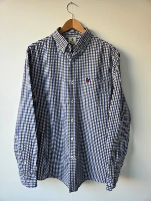 Blårutig skjorta från Lyle & Scott, storlek L - Klassisk blårutig skjorta från Lyle & Scott med vit och blå färg, långärmad modell och knappstängning. Skjortan har en bröstficka med broderad logga och är tillverkad i bomull. Perfekt för en avslappnad men stilren look. Size L.