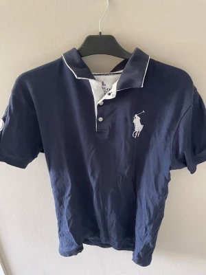 Mörkblå polo t-shirt från Ralph Lauren - Klassisk mörkblå polo t-shirt från Ralph Lauren med vit broderad logga på bröstet och vita detaljer vid krage och knäppning. Kortärmad modell i mjuk bomull, perfekt för en avslappnad och stilren look.