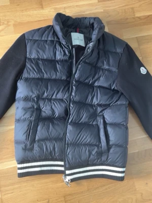 Svart cardigan från Moncler - Snygg svart cardigan från Moncler med quiltad front och bakstycke, samt stickade svarta ärmar. Västen har dragkedja framtill, två fickor och ribbade muddar med vita ränder. Klassisk Moncler-logga på ärmen. Perfekt för lager-på-lager och streetstyle.