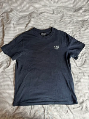 Mörkblå t-shirt från A.P.C. - Snygg mörkblå t-shirt från A.P.C. med broderad vit logga på bröstet. Klassisk rund hals och korta ärmar. Tillverkad i mjuk bomull och har en normal passform. Perfekt för en clean och stilren look.