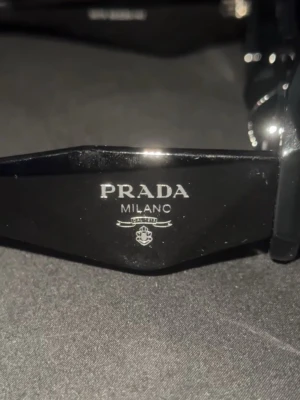 Prada solglasögon  - Snygga svarta solglasögon från Prada med klassisk och stilren design. Bågarna är i blank svart plast med tydlig Prada-logga på sidan. Perfekta för dig som vill ha en lyxig accessoar med italiensk känsla.