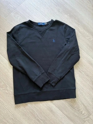 Svart sweatshirt från Polo Ralph Lauren - Klassisk svart sweatshirt från Polo Ralph Lauren med rund halsringning och blå broderad logga på bröstet. Tröjan har långa ärmar och ribbade muddar vid ärmslut och nederkant. Perfekt för en avslappnad och stilren look.