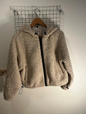 Beige teddyjacka med huva - Mysig beige teddyjacka med dragkedja framtill och stor huva. Jackan har två fickor på sidorna och justerbar dragsko nedtill. Perfekt för kalla dagar när du vill vara både varm och snygg. Tillverkad i fluffigt teddy-material💕💕