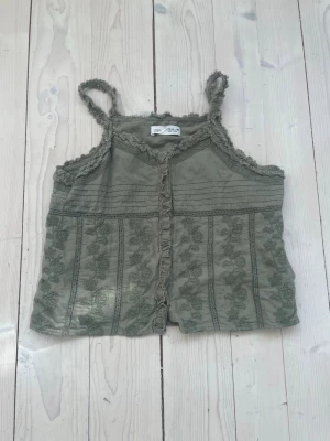 Topp från Zara - Snygg grön linnetopp från Zara med broderade blommor och spetsdetaljer längs kanterna. Toppen har smala axelband och knappar framtill. Perfekt för varma dagar och enkel att styla med jeans eller kjol.