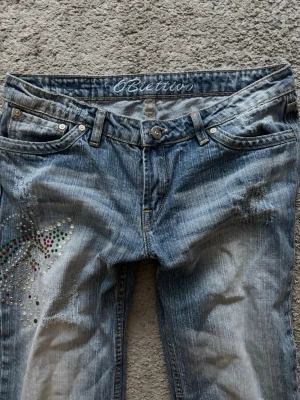 Blå jeans med strassdetaljer 6Blettivo - Snygga blå jeans från Obivetto med slitningar och coola strassdetaljer i färg på ena benet. Klassisk femficksmodell med raka ben och låg midja. Perfekta för dig som vill sticka ut lite extra med din denimstil.