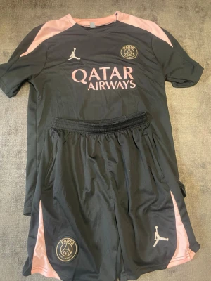 PSG träningsset svart/rosa Jordan - Snyggt PSG träningsset med t-shirt och shorts i svart med rosa detaljer. Setet har Paris Saint-Germain och Jordan-loggor samt Qatar Airways-tryck på bröstet. Tillverkat i lätt och ventilerande material, perfekt för fotboll eller träning.