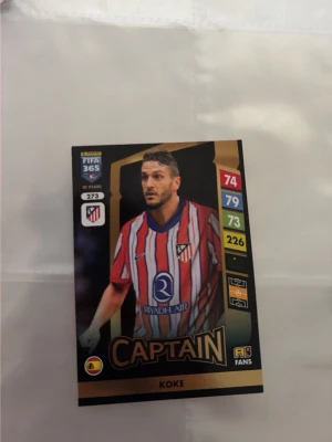Panini FIFA 365 fotbollskort - Två Panini FIFA 365 fotbollskort: ett med Koke från Atlético Madrid i rödvit randig tröja och ett med Miguel Borja från River Plate i vit tröja med röd diagonal. Båda korten har guldiga detaljer och spelarstatistik tryckt på framsidan.
