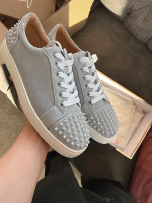 Gråa Louboutin sneakers med nitar - Snygga grå sneakers från Christian Louboutin med vita snören och ikonisk röd sula. Skorna har coola nitar på tån och hälen samt en stilren design i mocka. Perfekta för dig som vill sticka ut med exklusiva detaljer och streetstyle-vibe.