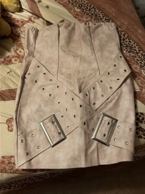Beige axelbandslös klänning med bälte - Trendig beige axelbandslös klänning med två breda bälten med silverspännen framtill. Klänningen har en kort passform och är tillverkad i ett mjukt material med mockakänsla. Perfekt för dig som vill sticka ut med en edgy look.