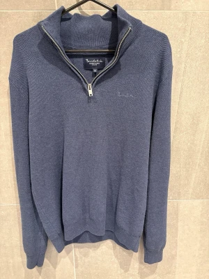 Blå quarterzip från Bondelid - Mörkblå stickad tröja från Bondelid med halv dragkedja och hög krage. Tröjan har ribbade muddar vid ärmslut och nederkant, samt diskret broderad logga på bröstet. Perfekt för lager-på-lager och snygg till jeans.