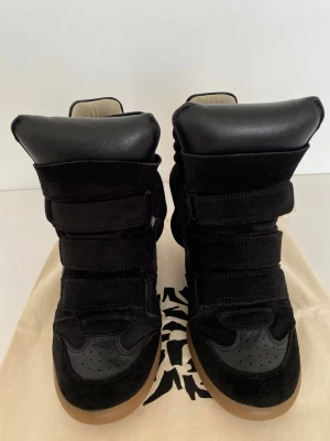 Svarta sneakers från Isabel Marant - Säljer ett par svarta high-top sneakers från Isabel Marant med cool design. Skorna har breda kardborreband, detaljer i mocka och skinn samt en klassisk gummisula i beige. Perfekta för dig som vill ha en statement sneaker med unik look.