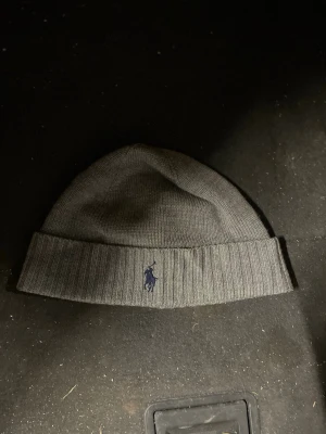 Grå mössa från Polo Ralph Lauren - Snygg grå ribbstickad mössa från Polo Ralph Lauren med klassisk broderad logga framtill. Perfekt för kalla dagar och enkel att matcha med olika outfits. Mjuk och bekväm passform.