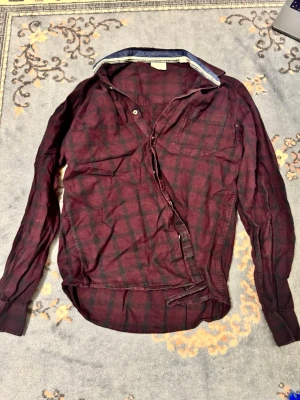 Vinröd rutig skjorta med knappar - Vinröd och svart rutig skjorta med lång ärm och klassisk krage. Skjortan har knappar framtill och en bröstficka. Tillverkad i ett mjukt och lätt material, perfekt för en avslappnad stil.