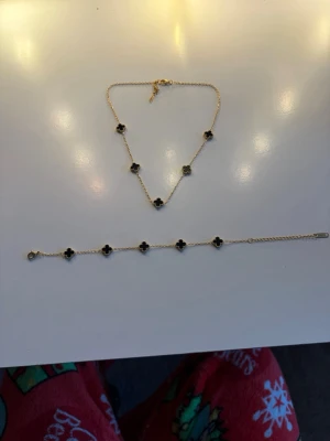 Guldhalsband och armband med svarta klöverhängen - Snyggt halsband och armband  i guldton med fem svarta klöverformade hängen. Kedjan är tunn och elegant, perfekt för att ge en stilren touch till din outfit. Klöverhänget ger en trendig och lyxig känsla. Passar dig som gillar klassiska smycken med en twist.