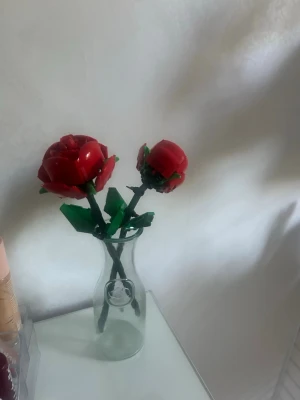 Superfina Lego rosor  - Två röda rosor byggda av Lego med gröna blad och stjälkar, placerade i en genomskinlig glasvas. Perfekt som dekoration till skrivbordet eller hyllan. Rosorna är detaljerade och har en cool, modern känsla.