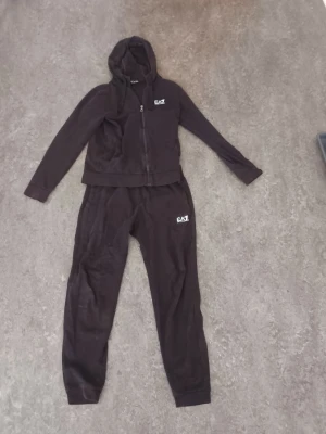 Svart EA7 loungewear-set med hoodie - Säljer ett svart loungewear-set från EA7 Emporio Armani. Setet består av en zip-hoodie med huva och matchande byxor, båda med vit EA7-logga. Perfekt för chill dagar hemma eller när du vill vara bekväm men ändå snygg.