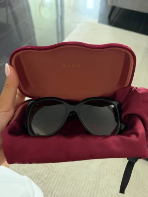 Svarta Gucci solglasögon i cat eye - Snygga svarta solglasögon från Gucci med cat eye-formade bågar. Glasögonen har breda skalmar med Gucci-logga i guld och diskreta ränder i rött och grönt på sidorna. Kommer med originalfodral i vinröd sammet.