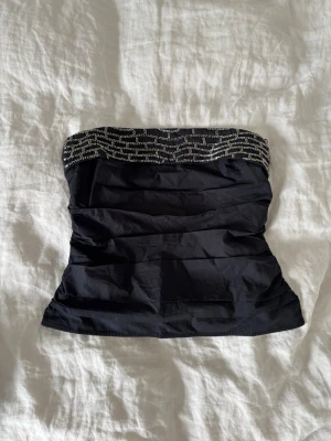 Svart tubtopp med silverdetaljer - Snygg svart tubtopp med glittriga silverdetaljer upptill och dragkedja i ryggen. Toppen är figurnära och axelbandslös, perfekt för att skapa en cool och trendig look.