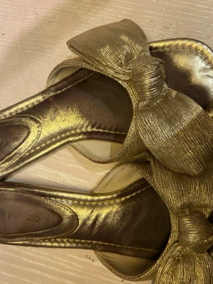 Guldiga sandaler med stor rosett NA-KD - Snygga sandaler i metallic guld från NA-KD med bred rem och stor rosett framtill. 