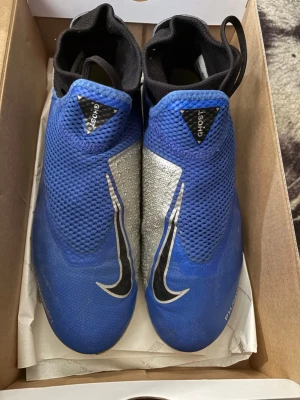 Nike Phantom Vision blå fotbollsskor - Säljer ett par Nike Phantom Vision fotbollsskor i blått och silver med svarta detaljer. Skorna har en strumpliknande och en stor Nike Swoosh på sidan. Ovandelen är i syntetmaterial med struktur för bättre bollkontroll. Perfekta för dig som vill sticka ut på planen. Skorna passar även 42,5