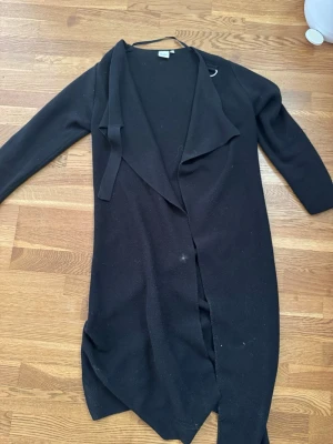 Svart lång cardigan med sjalkrage - Svart lång cardigan med öppen front och sjalkrage. Modellen är rak och har långa ärmar. Perfekt att slänga över en topp för en avslappnad look. Materialet är mjukt och stickat, vilket gör den skön att bära.