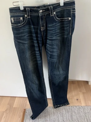 Mörkblå Miss Me jeans med kontrastsömmar - Snygga mörkblå jeans från Miss Me med vita kontrastsömmar och dekorativa bakfickor med broderier och nitar. Jeansen har klassisk femficksdesign, mid rise och raka ben. Perfekta för dig som gillar detaljer och en cool look.